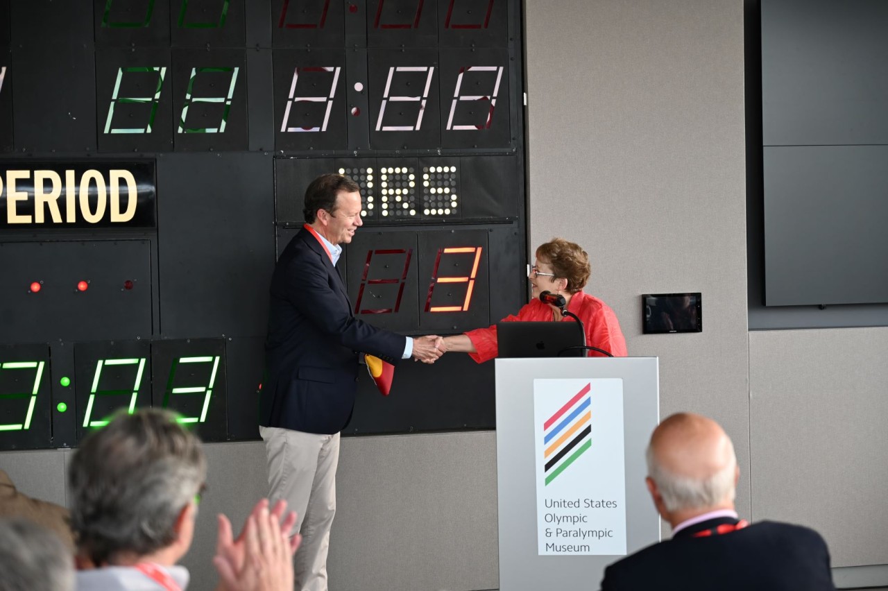 Representative Terri Carver shaking hands with and presenting the Colorado state flag to WACA Board of Directors Chairman Glenn Creamer in front of the “Miracle on Ice” scoreboard.